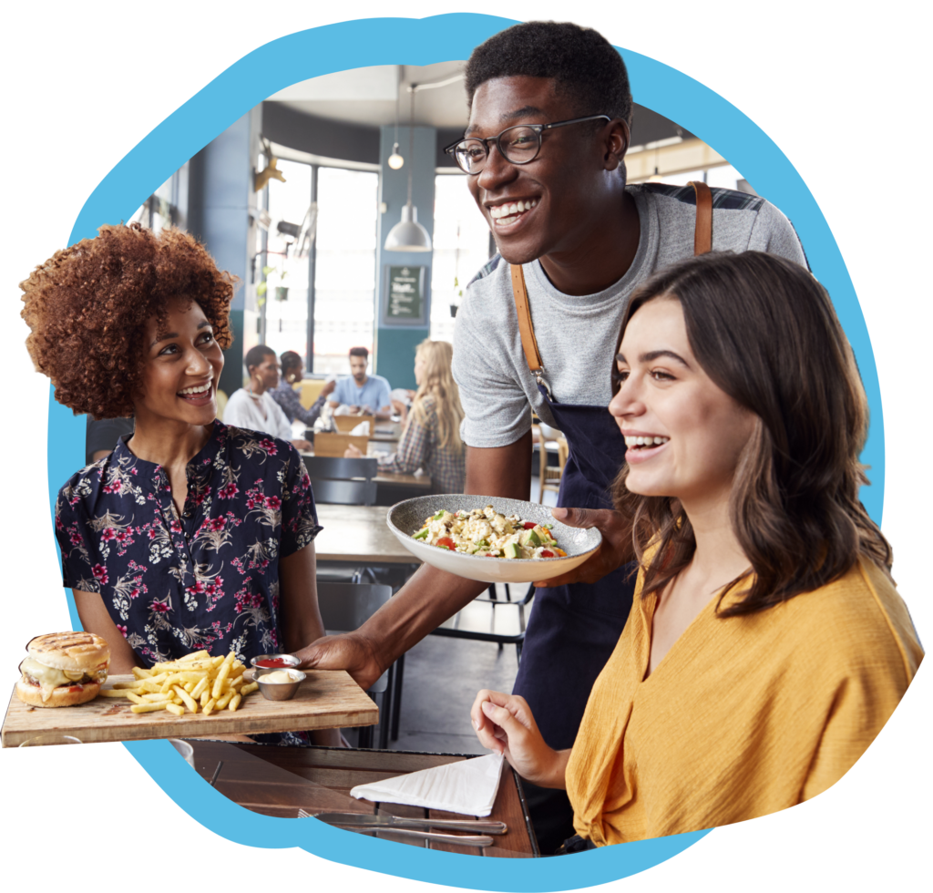 A young happy African American male server delivers hamburger and french fries and a chopped salad to an African American female and her caucasian female friend. 