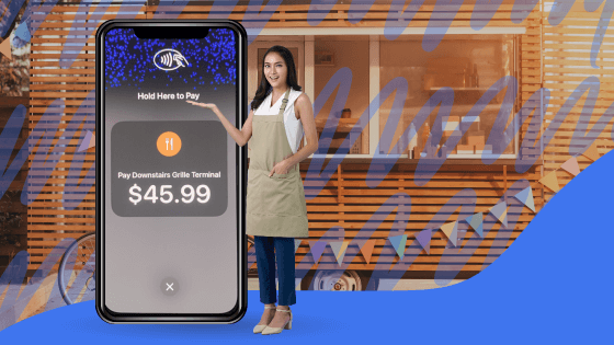 A young female food truck operator smiles while standing beside an oversized smartphone displaying Table Needs' Mobile App. She demonstrates how to accept payments with a tap-to-pay feature.