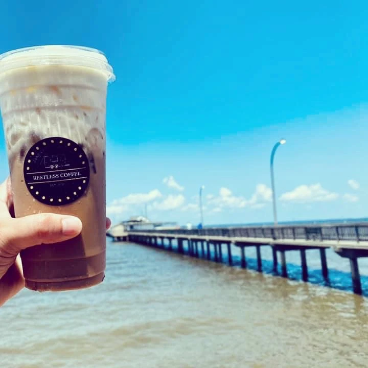 The image features a person's hand holding up a cold beverage from Restless Coffee against a coastal backdrop. The drink is in a clear cup with the company's logo prominently displayed, and the setting includes a bright blue sky, a calm sea, and a long pier extending into the water, evoking a sense of relaxation and leisure. This picture conveys the idea of enjoying a refreshing Restless Coffee drink while taking in the serene views of a seaside environment.
