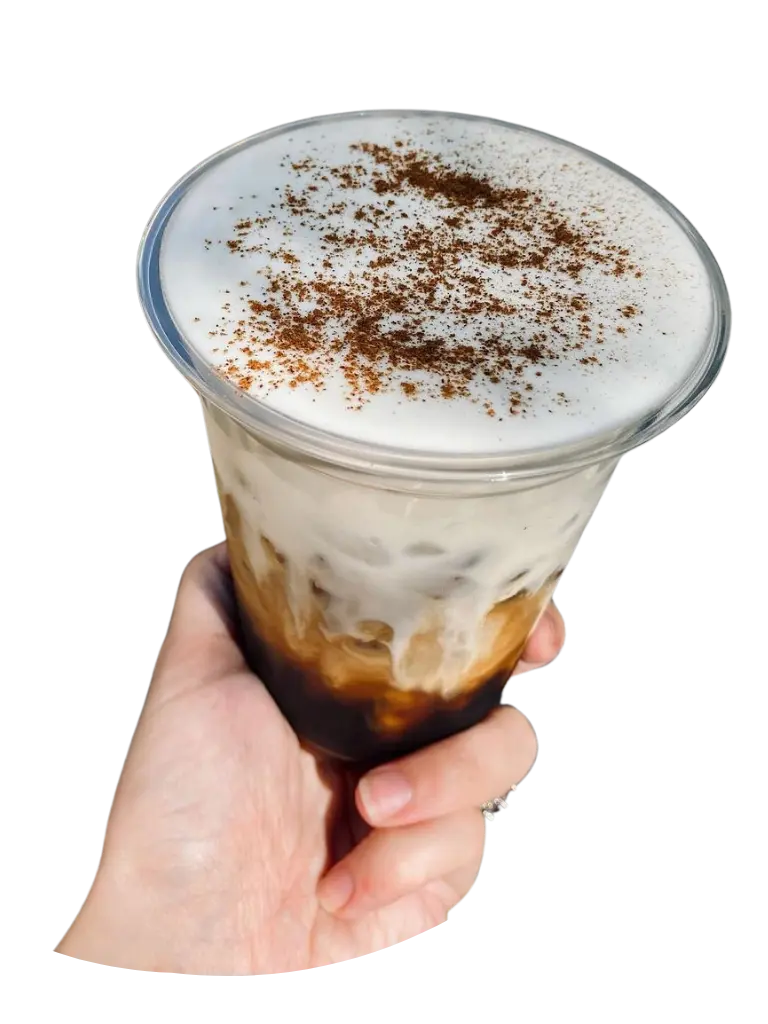 The image captures a hand holding a beautifully crafted coffee drink with a frothy, creamy top lightly dusted with a sprinkle of brown spice, likely cinnamon or cocoa, contrasting against the darker coffee below. The drink exemplifies the handcrafted quality that Restless Coffee is known for, representing the meticulous preparation that goes into each order, allowing them to start fulfilling customer demands promptly, as Christian mentions in his quote about the satisfaction and efficiency of having orders ready before opening.