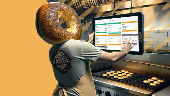 A person with a bagel for a head stands in a kitchen, using a touchscreen kitchen display system. The screen shows a colorful interface with various orders and instructions. In the background, freshly baked bagels are visible on the counter.