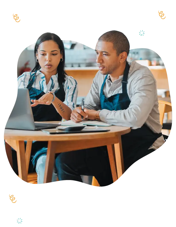 Two restaurant team members, a man and a woman wearing aprons, are engaged in a focused business discussion at a wooden table. They are surrounded by a casual dining setting, with the woman pointing to something on a laptop screen while holding a pen, suggesting they are analyzing data or scheduling. The image is encapsulated in a unique cut-out design with decorative elements like utensils and a coffee cup, indicating a hospitable and food-related work environment. This setting underscores the image's relevance to independently-owned quick service restaurants, bakeries, cafes, and food trucks, highlighting technology and business services for profitability.